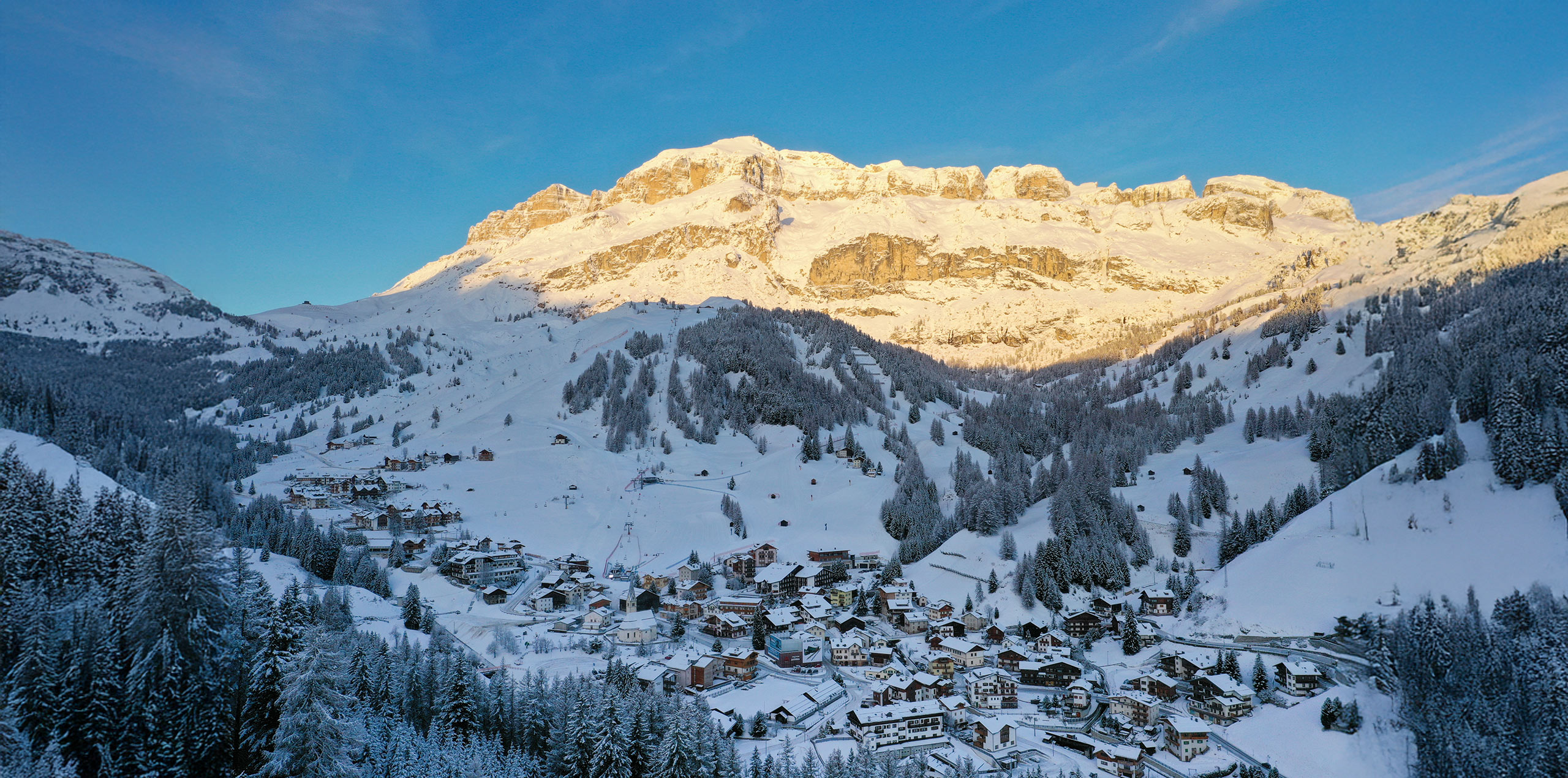Arabba, heart of the Dolomites, on a winter morning. The snow-covered village is surrounded by fir trees and sunlit mountains.