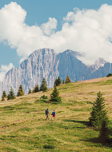Two hikers walk on a grassy mountain path, with the Dolomites and white clouds in the background.