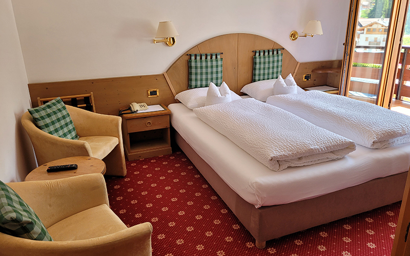 Gemütliches Doppelzimmer im Garni Royal in Arabba mit Einzelbetten, Holzmöbeln, Sesseln und Balkon mit Blick auf die Dolomiten.