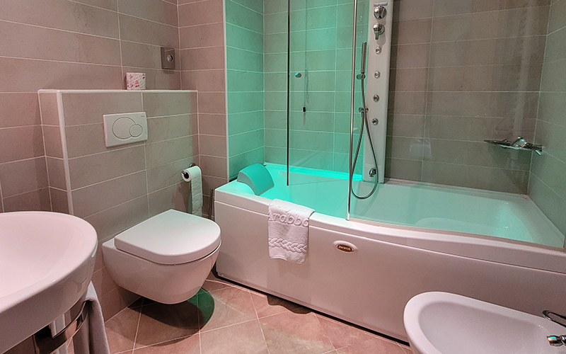 Modernes Badezimmer mit hellen Fliesen, grün beleuchteter Whirlpool-Badewanne, Wanddusche und hängenden Sanitäranlagen.