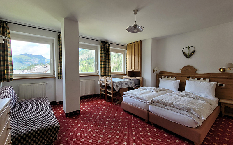 Spacious mountain-style bedroom at Apartments Alpenroyal in Arabba, with single beds, a sofa, and a view of the Dolomites.