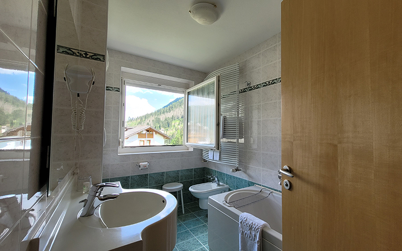 Bright bathroom of an Alpenroyal apartment with an open window to the Dolomites, a washbasin, bidet, and bathtub.