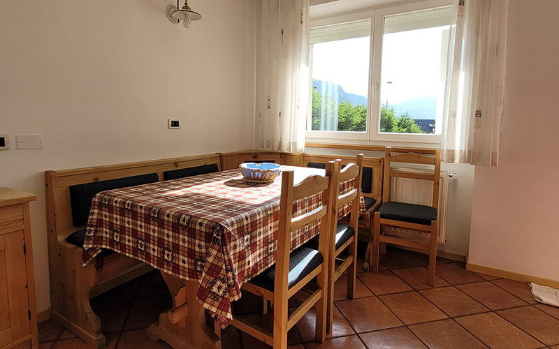 Light wood dining area with bench and chairs, table with checkered tablecloth and view of the Dolomites.