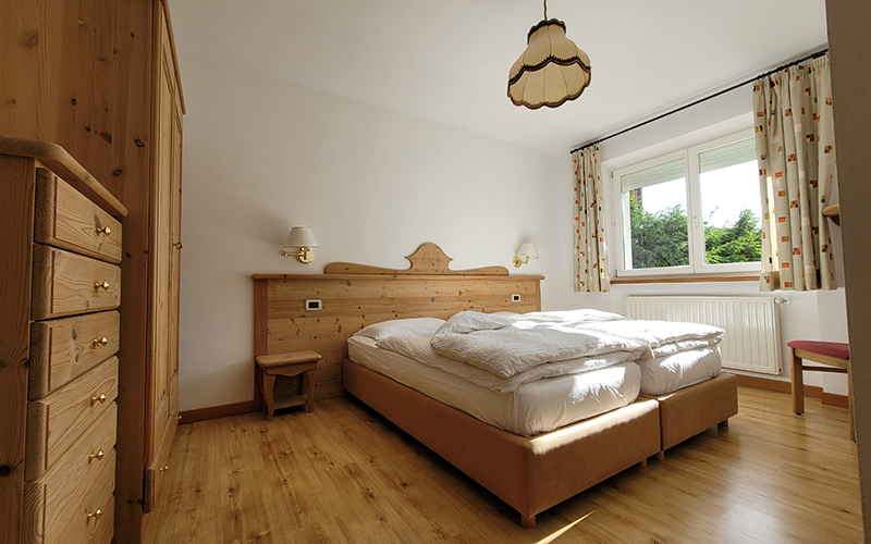Mountain-style double bedroom with light wood furniture, bed with white duvets, and window with a view of the Dolomites.