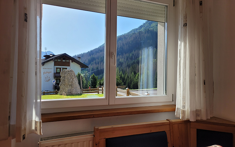 Window of an Alpenroyal apartment with a view of a mountain-style building and the Dolomites of Arabba.