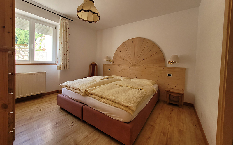 Double bedroom in alpine style of the Appartamento Alpenroyal in Arabba, with bed, wooden headboard, and window with Dolomites view.