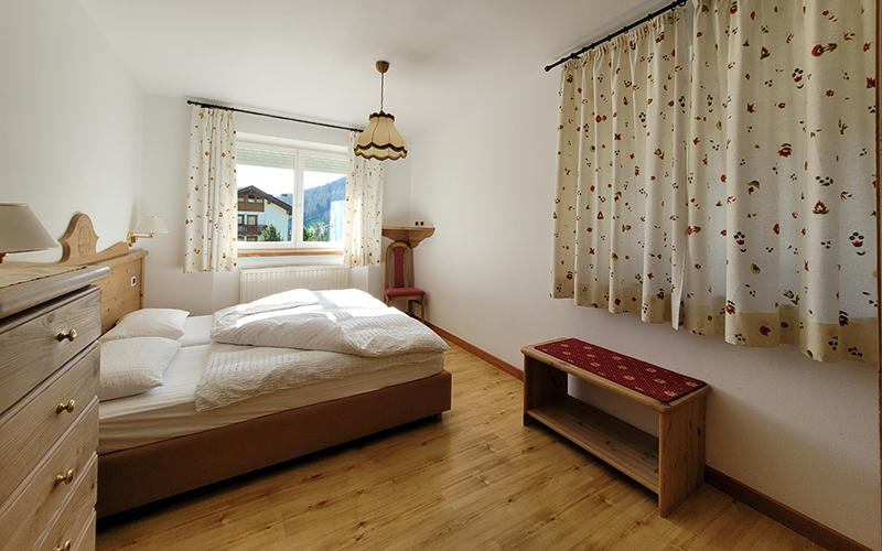 Double bedroom in alpine style of the Alpenroyal Apartment, with bed, nightstand, and window with a view of the Dolomites of Arabba.