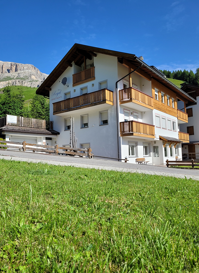 Appartamenti Alpenroyal in Arabba: White building with wooden balconies and Dolomite mountains in the background.