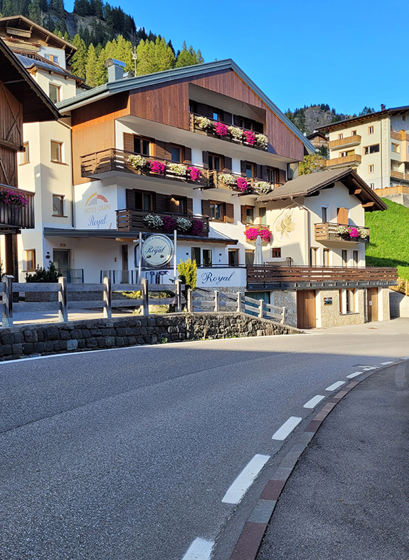 The Hotel Garni Royal in Arabba, Dolomites, with flowered balconies and green mountains in the background, on a sunny day.