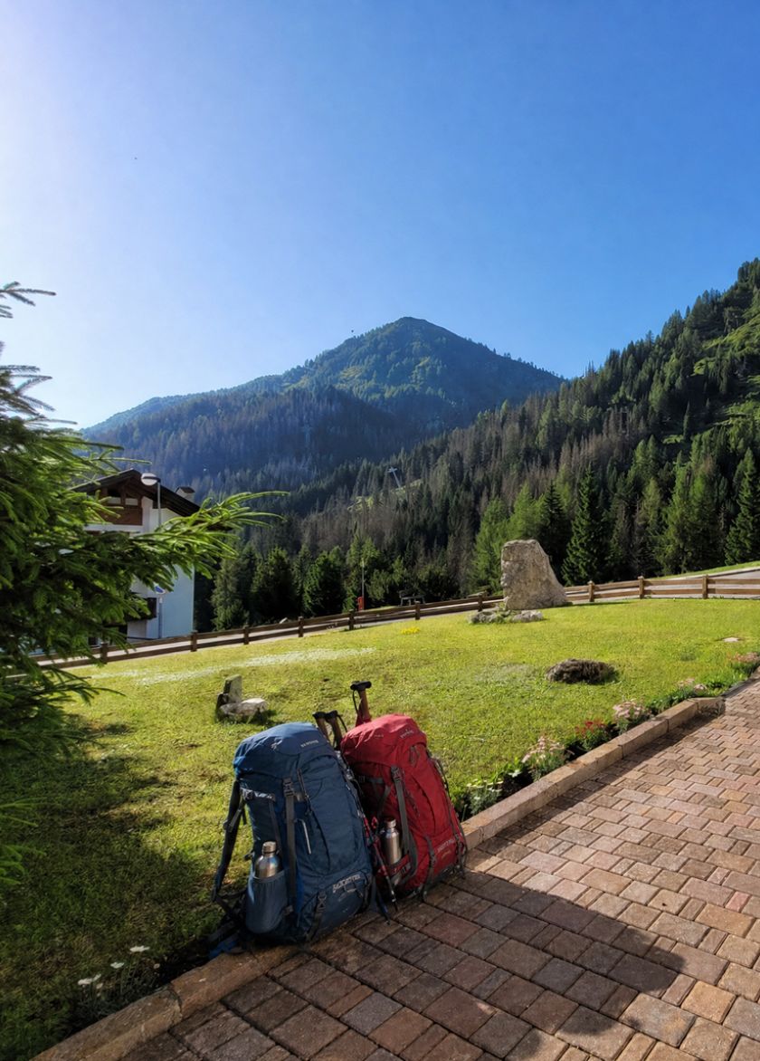 Two blue and red trekking backpacks resting on a stone path, with the Dolomites and forest in the background.