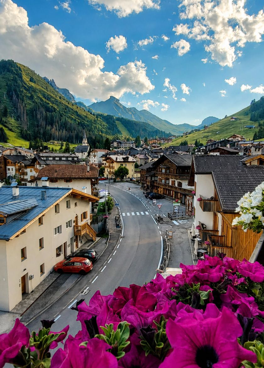 Panoramablick auf Arabba im Sommer, mit typischen Häusern, einer sich durch die grünen Berge der Dolomiten schlängelnden Straße und fuchsiafarbenen Blumen im Vordergrund.