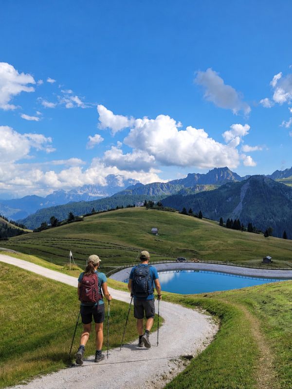 Couple trekking in the Dolomites near Arabba, with backpacks and trekking poles, on a dirt path alongside an alpine lake.