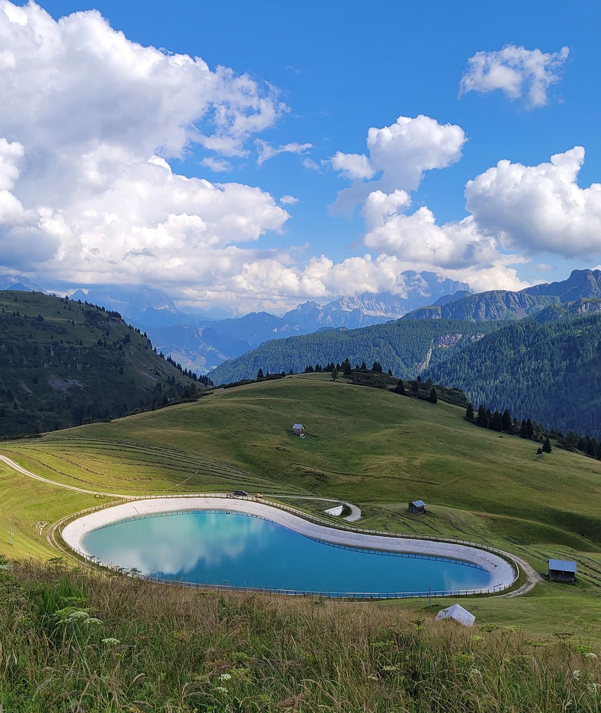 Vista panoramica estiva delle Dolomiti ad Arabba con un lago artificiale turchese e nuvole riflesse.