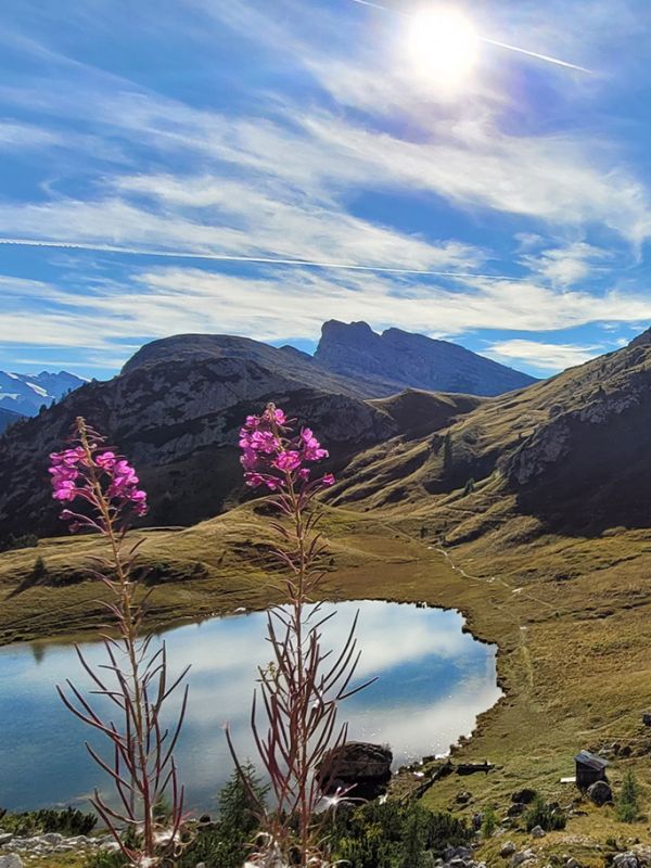 Paar beim Trekking in den Dolomiten bei Arabba, mit Rucksäcken und Wanderstöcken, auf einem Schotterweg entlang eines Alpensees.
