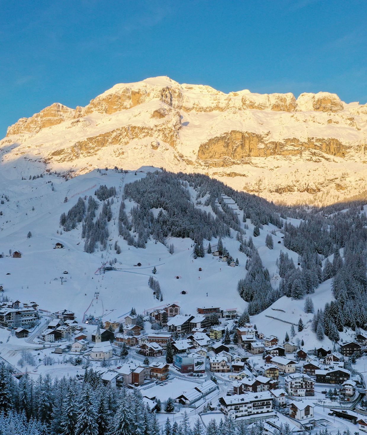 Arabba, cuore delle Dolomiti, in un mattino d'inverno. Il paese innevato è circondato da abeti e montagne illuminate dal sole.