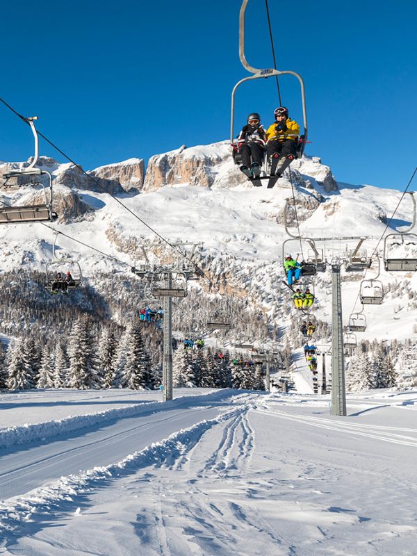 Skiers ascend by chairlift on the snow-covered Dolomites of Arabba, with slopes and forests under a blue sky.