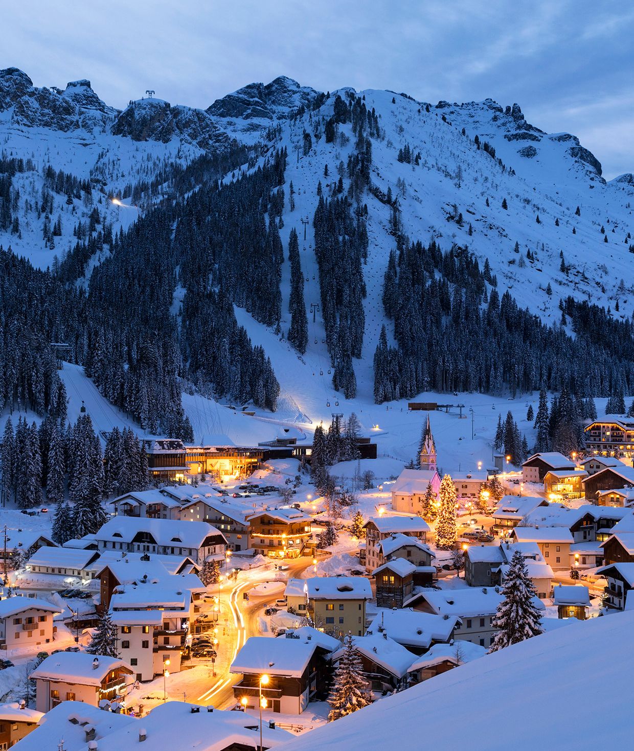 Panorama notturno di Arabba innevata, con case illuminate, piste da sci e impianti di risalita.