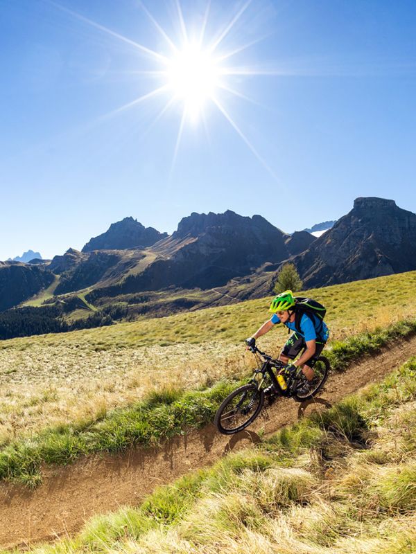 A mountain biker on a dirt path through grass, with the Dolomites and bright sunshine in the background.