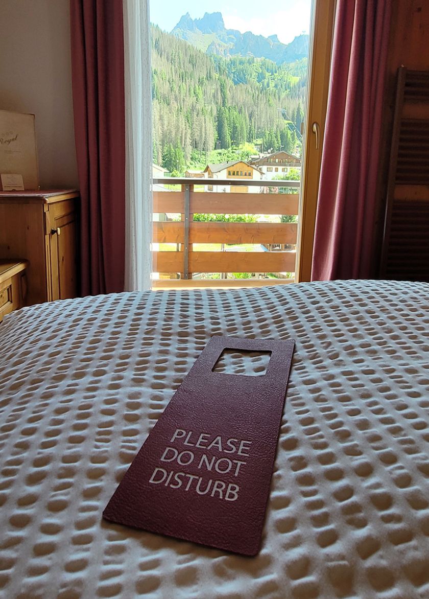 Zimmer des Garni Royal in Arabba mit einem „Bitte nicht stören“-Schild auf dem Bett und Panoramablick auf die Dolomiten.