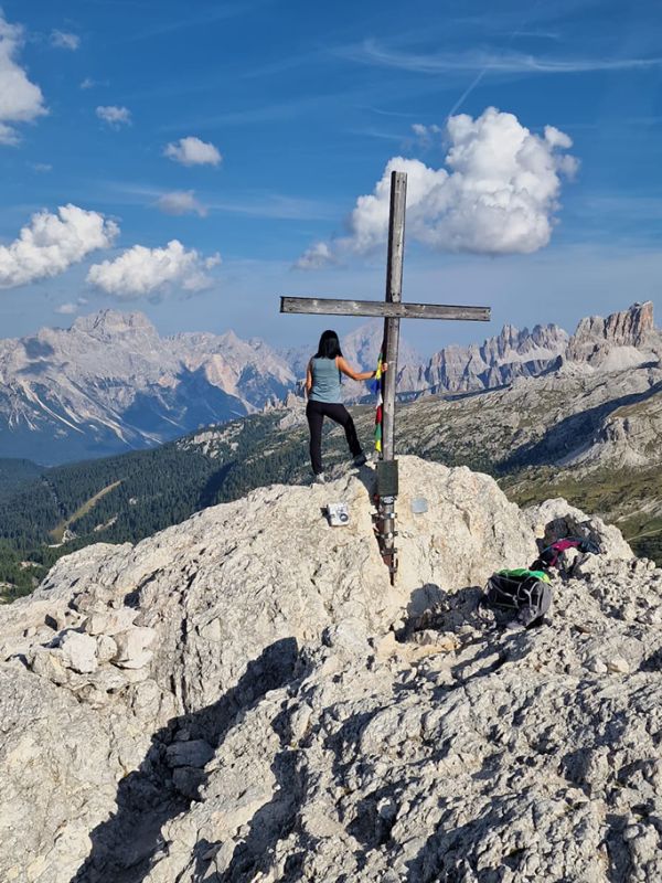 Hiker from behind on a rocky summit with a cross, admiring the Dolomites of Arabba under a blue sky.