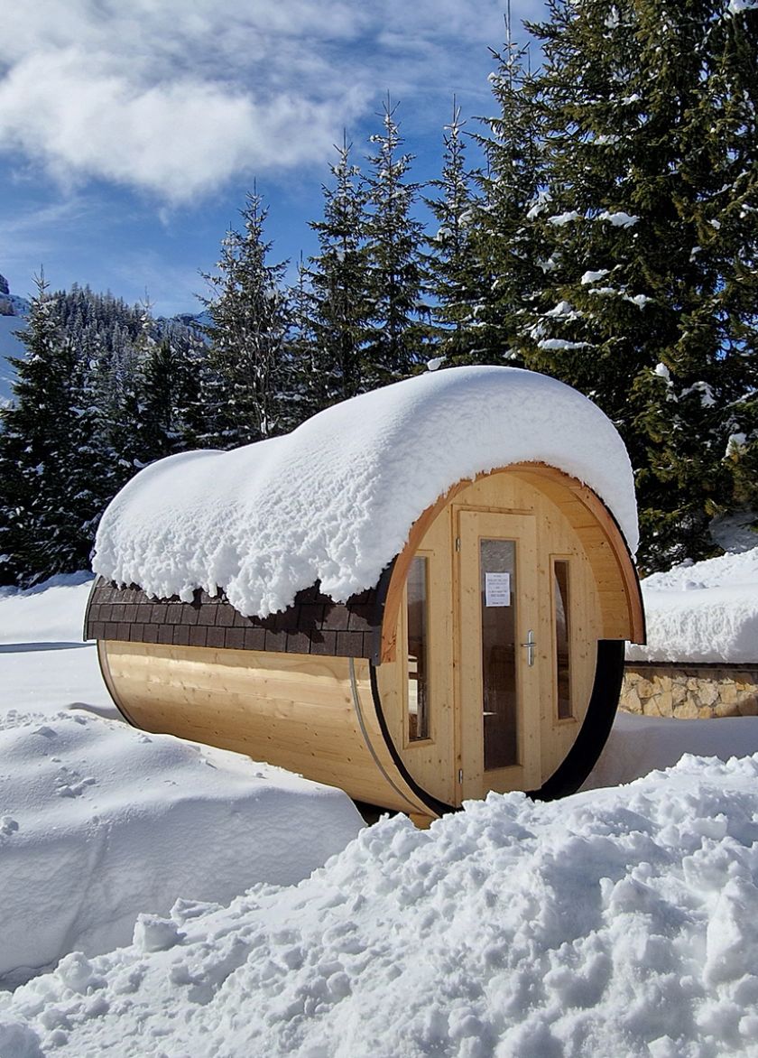 A wooden barrel sauna, covered in fresh snow, nestled in the winter landscape of the Dolomites.