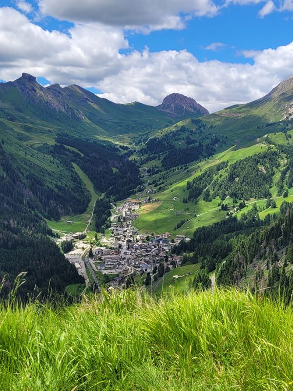 Panoramablick auf Arabba und das Tal, umgeben von den Dolomiten, mit grünen Wiesen im Vordergrund.